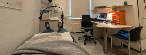 Ein Mann liegt in einem Behandlungsraum auf einer Liege. Man sieht den Köroer ab der Hüfte abwärts. im Hintergrund ein Ständer für Infusionen, an der Wand rechts ein Schreibtisch mit Stuhl und einem Computer auf der Tischplatte.