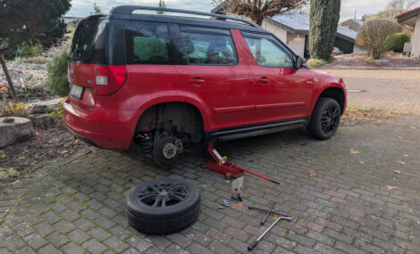Ein roter Skoda Yeti steht auf einer gepflasterten Fläche. Er ist hinten mit einem kleinen Hydraulilrangierwagenheber hoch gebockt und das rechte Hinterrad liegt demontiert neben dem Fahrzeug. Es liegen auch ein paar Radmuttern und entsprechendes Werkzeug herum.