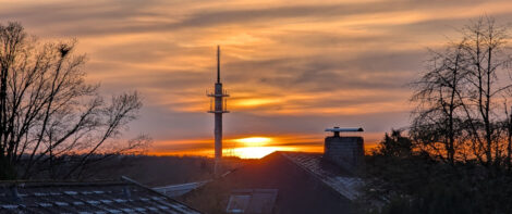 Die untergehende Sonne färbt den Himmel goldgelb. Man sieht einige Dächer von Häusern und Baumspitzen. In der Mitte des Bildes direkt neben der am Horizont stehenden Sonne sieht man einen Postfunksendemast.