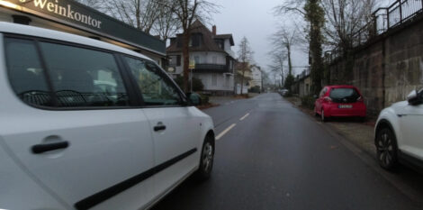 Ein Foto aus der Lenkerperspektive eines Fahrrades. Rechts parken KFZ in Parkbuchten. Das Fahrrad wird mit ca. 1 Meter Abstand zu den parkenden KFZ gefahren. Links sieht man ein überholendes KFZ, welche mit der rechten Spur die eingezeichnete Mittellinie deutlich nicht überfahren hat und somit einen äußerst knappen Abstand zum Menschen auf dem Fahrrad einhält.