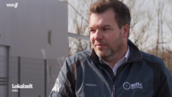 Ein Screenshot aus der Lokalzeit OWL. Man sieht einen Mann (mich) in einer ADFC-Jacke von Vaude. Im Hintergrund ist ein Teil einer Halle und kahle Bäume zu sehen.