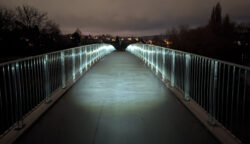 Mittiger Blick über eine gebogene Brücke mit von einer Fahrradlampe beeuchtetem Geländer rechts und links. Es ist dunkel und im Hintergrund erkennt man eine beleuchtete Häuserzeile.