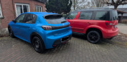 Von schräg links hinten fotografiert: ein blauer Peugeot E 208 GT und rechts daneben ein roter Skoda Yeti Monte Carlo vor einem Einfamilienhaus mit Garten.