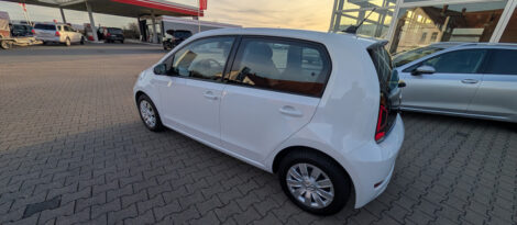 Ein weißer Volkswagen e-Up steht auf dem großen Hof eines Autohauses. Im Hintergrund sieht man eine Tankstelle.