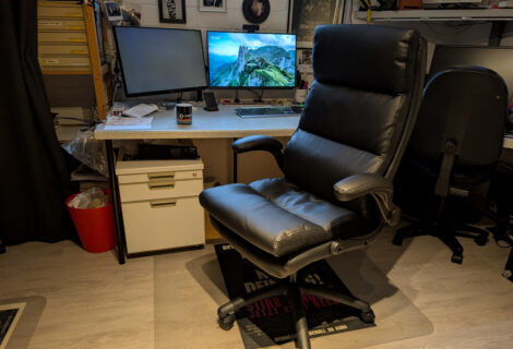 Ein schwarzer Kunstleder-Bürostuhl vor einem Schreibtisch mit zwei 27Zoll-Monitoren. Unter dem Schreibtisch ist ein Rollcontainer zu sehen.