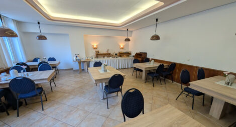 Sachlich eingerichteter Frühstücksraum eines Hotels. Beide Bodenfliesen, wuchtige '80er-Jahre Tische und blaue Stühle.