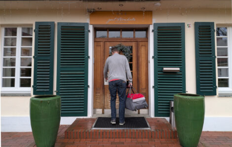 Ein Mann steht mit dem Rücken zur Kamera vor einer Eingangstür zu einem Haus. Rechts und links der Tür sieht man grüne Lamellengitter, zum Abdecken von Türen und Fenstern.