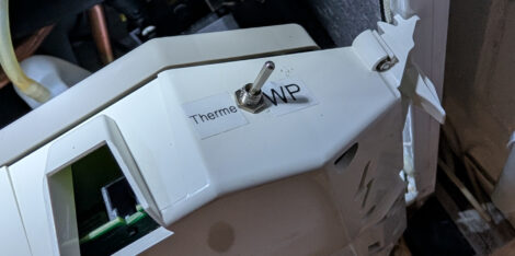 In einer weißen Plastikverkleidung ist ein kleiner, verchromter Kipschalter montiert. Auf der einen Seite des Schalters ist "WP" neben dem Schalter zu lesen, auf der anderen Seite "Therme".