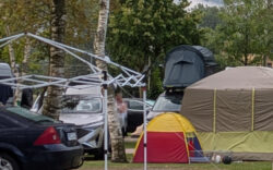 Ein Campingplatz. Man sieht im Vordergrund das Gestell eines Faltpavillons ohne die Zeltstoff und im Hintergrund, hinter einigen Bodenzelten sieht man ein grünes Dachzelt auf einem Van.