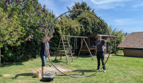 Zwei Männer stehen auf einer grünen Rasenfläche und halten einen Kreis aus Metallrohren senkrecht in die Luft. Im Hintergrund steht eine Kinderschaukel aus Holz und es sind hohe Kirschlorbeerhecken zu sehen.