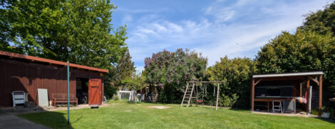 Ein grüne Rasenfläche. Links sieht man einen rotbraunen Carport mit integriertem Geräteschuppen, ganz rechts einen Unterstand, in dem ein Dachzelt aufgebaut ist. In der Mitte dazwischen steht eine Kinderschaukel aus Holz. Der Carport wird überspannt von den Ästen zweier großer Buchen, der restliche Garten ist von hohen Hecken und Büschen umgeben.