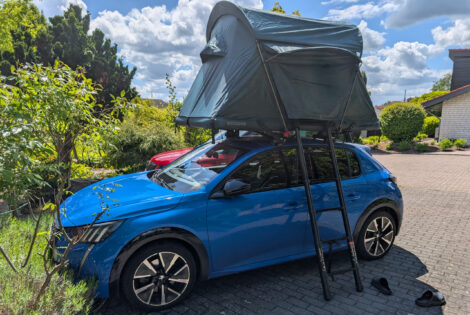 Ein blauer Peudeot E 208 GT steht auf einem Parkplatz mit einem Garten im Hintergrund. Auf dem Dach des Peugeot ist ein grünes Dachzelt "Groenberg Weekend Comfort" montiert. Man schaut von schräg vorne auf die Fahrerseite des Fahrzeugs. An die Seite des Dachzeltes ist eine Ausziehleiter angelehnt. Am eigentlich blauen Himmel ziehen reichlich weiße Wolken vorbei.