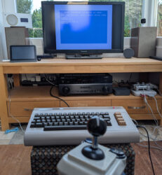 Auf einem Sideboard steht ein Fernseher auf dem der Einschaltbildschirm eines C64 zu sehen ist. Links und rechts des Fernsehers stehen zwei alte Telefunken-Boxen, welche von dem im Fach unter dem Fernseher platzierten Yamaha AX570 angetrieben werden. Im Vordergrund steht auf einem Hocker ein beiger C64 und davor ein ebenso beiger ArcadeR-Joystick.