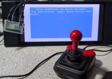 Ein kleiner Raspberry Pi hängt über einem kleinen Widescreen-TFT, auf dem Einschaltbild eines C64 zu sehen ist. Davor sieht man einen "Competition Pro"-Joystick.