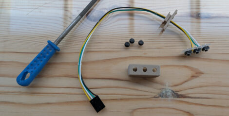 Ein mehradriges Kabel an dessen Ende eine kleine Platine mit drei Tastern montiert ist. Daneben liegt ein dazu passendes beiges Gehäuse, eine Grundplatte ist am Kabel belassen und drei sehr kleine Taster. Weiterhin ist eine Reibahle mit einem blauen Griff zu sehen.