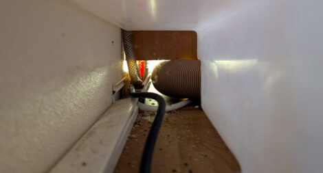 Der Bick durch einen Tunnel hinter dem Wassertank eines Wohnwagens in der Sitzbank desselben. Man sieht einen Kabelkanal und an dessen Ende einige Kabel und Wasserschläuche, die dahinter in einen Schrank führen.