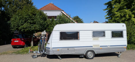 Ein Wohnwagen steht auf einer gepflasterten Fläche vor einem Einfamilienhaus in dessen Vorgarten Büsche und Bäume wachsen.