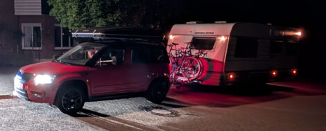 Ein Wohnwagengespann steht nachts auf einer gepflasterten Fläche. Beim roten Zugfahrzeug sind die Frontscheinwerfer an und auch die Positionsleuchten des Wohnwagen brennen.