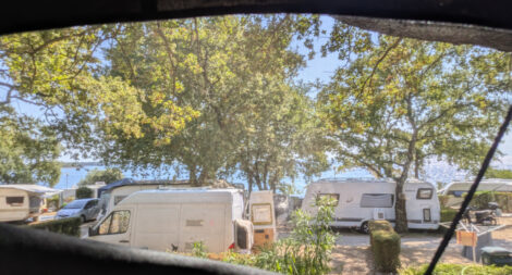 Blick aus dem Fenster eines Dachzeltes durch Bäume und über Wohnwagen hinweg auf das Meer der Adria.