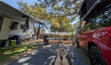 Ein Wohnwagenstellplatz. Vor einem Caravan mit Markise stehen Campingmöbel. REchts im Bild ist das Zugfahrzeug mit einem aufeklappten Dachzelt.