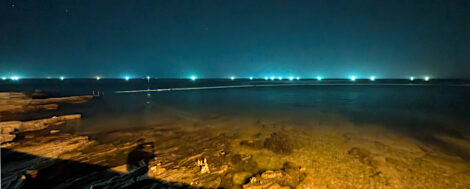 Die Adria bei Nacht. Die Laternen der Strandpromenade beleuchten das sehr flache Wasser der Küste und in der Ferne erhellen die Positionslichter der Fischerboote den Horizont.