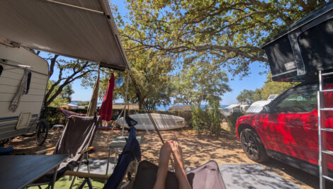 Zwei Füße ragen aus einer Hängematte. Links im Bild ein Wohnwagen mit einer Markise, rechts ein Auto mit einem Dachzelt. Im Hintergrund eine Hecke und dahinter sieht man über weitere Wohnwagendächer das Meer.