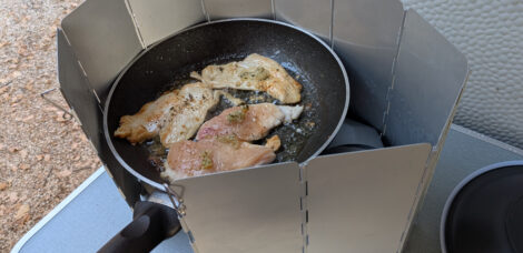 Auf einem CampingGaz-Kocher ist eine Pfanne mit Hähnchenchnitzeln. Um den Kocher ist ein Aluminium-Windschutz aufgebaut.