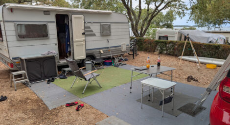 Ein Wohnwagen mit davor ausgelegten Vorzeltteppichen und eingefahrener Sackmarkise Auf den Teppichen stehen noch einige Campingmöbel.