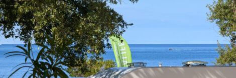 Blick auf das Meer mit einem Baum links im Bild. Man blickt über Caravandächer auf die Adria und mitten im Bild sieht man den oberen Teil einer Herbalife-Beachflag.
