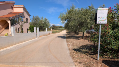 Eine schmal asphaltierte Straße. Links sieht man mediterane Einfamilienhäuser, rechts kleine Bäume. Im Vordergrund steht ein Hinweis auf den Parenzana-Radweg.