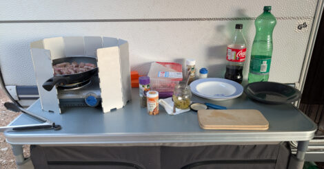 Kochuntensilien auf einem faltbaren Campingschrank. Auf einem Gaskocher mit aufgebautem Windschutz steht eine Pfanne mit Hackröllchen. Auf dem Regal daneben allerlei Öl und Gewürze.