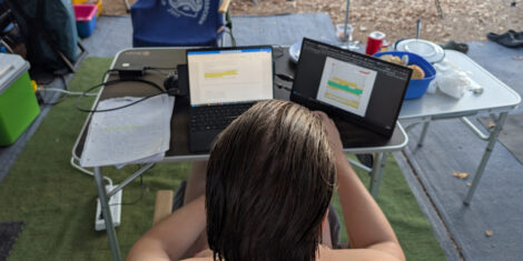 Der Hinterkopf eine jungen Mannes vor einem Laptop mit Zusatzmonitor. Das Equeipment ist auf einem Campingtisch aufgebaut.
