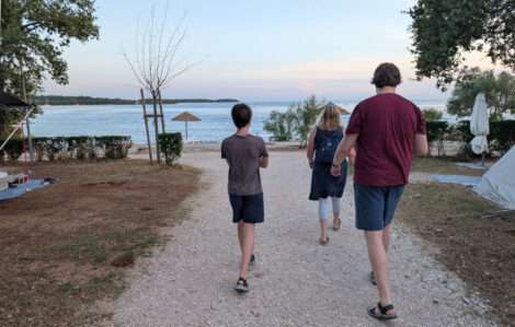 Drei Menschen gehen auf einem asphaltierten Weg auf das Meer zu.