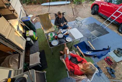 Ein junger Mann und eine Frau sitzen unter einer Markise an einem gedeckten Campingtisch. Die Kamera blickt von oben auf die Szene.