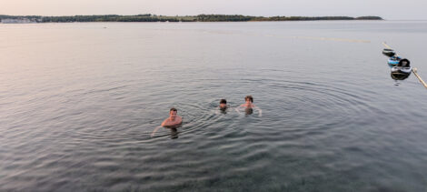 Ein Papa, ein älterer und ein jüngerer Sohn baden in der Adria. Es schauen nur die Köpfe aus dem Wasser, welches völlig glatt ist. Im Hintergrund sieht man die Landzunge auf der anderen Seite der Bucht. Rechts dümpeln ein paar StandUp-Paddelbretter, die an einer Leine zur Abgrenzung des Schwimmbereichs befestigt sind.