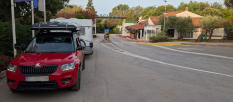 Ein rotes Auto mit Dachzelt steht vor einem Wohnwagen. Im Hintergrund sieht man die Einfahrt zu einem Campingplatz mit einem großen Schriftzug "Solaris" über den Schranken.