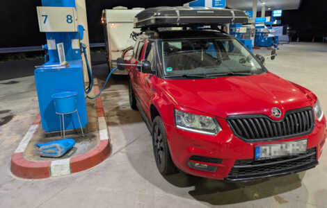 Ein rotes Auto mit einem zusammengeklappten Dachzelt oben drauf steht vor einem Wohnwagen an der Zapfsäule einer Aral Tankstelle.