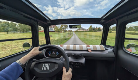 Foto nach vorne aus einem Fiat Topolino. Durch das Glasdach sieht man den blauen Himmel mit kleinen weißen Wolken. Am Lenkrad die Hände einer Frau.