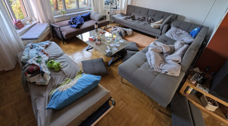 Ein Wohnzimmer mit drei Sofas, einer Liege und einem Luftbett. Es liegen Schlafutensilien und Schlickersachen herum.